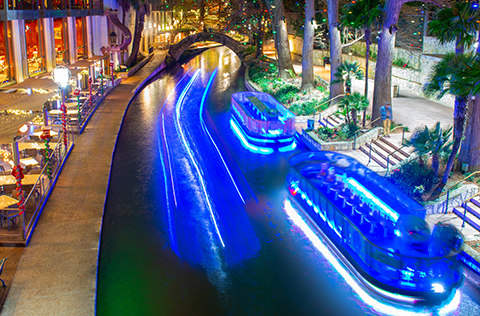 Two boats parked on the side of the River Walk, with another boat passing.