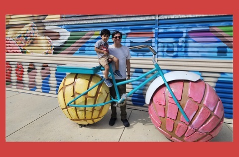 A man holding his son standing next to a bike with concha wheels and a colorful mural.