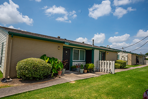 Cottage Creek Apartments are one story with white siding, green trim including a frontal beige brick wall and some landscaping.