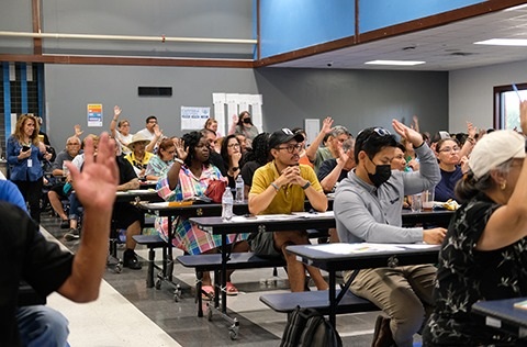 Community workshop with attendees seated at tables raising their hands in a meeting room.