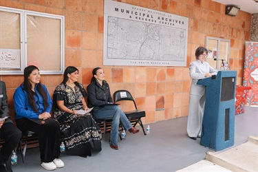 City of San Antonio City Clerk standing at the podium while the next speakers sit nearby, preparing to speak at the Grand Opening.