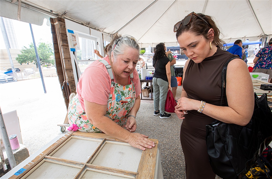 A woman observes another woman examining a window frame at a home repair fair, surrounded by various home improvement displays.