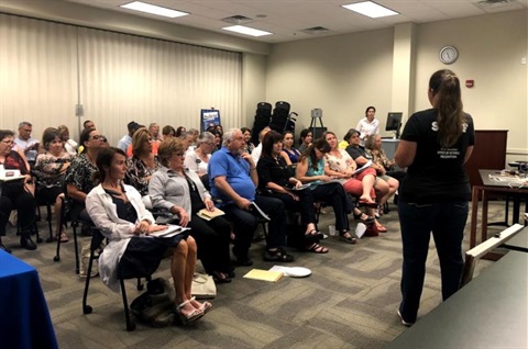 A staff member presenting to a seated audience during a meeting in a conference room setting. 