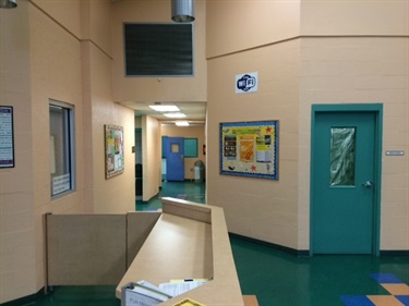 Front desk and bulletin board on the side wall. Hallway area behind shows doorways to several rooms.