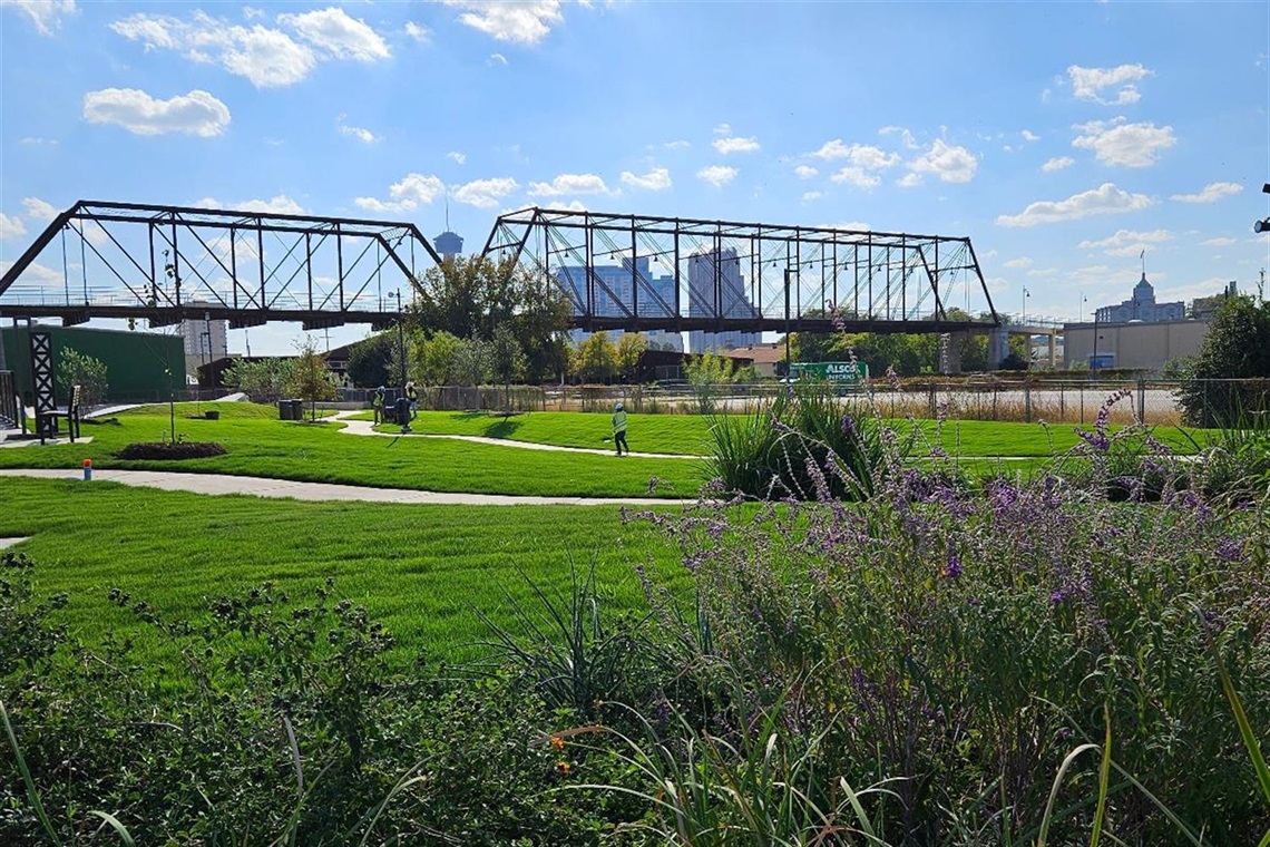 A bridge surrounded by green landscape with the City of San Antonio skyline in the background.