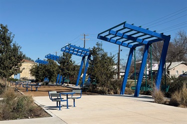 Park seating area with blue shade structures, picnic tables and plants along a paved walkway