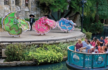 People on a blue boat in a river, watching a Mexican folk dance with a woman in a colorful Jalisco Ribbon dress.