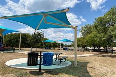 In the middle of a park, a blue umbrella offers shade over picnic tables, close to trash and recycling cans.