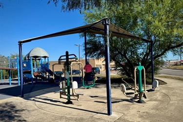 Outdoor gym equipment beside a playground, with a person exercising under a shade canopy.