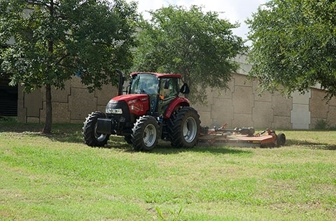 A red tractor with a mowing attachment drives through a lush grassy field under a clear blue sky.