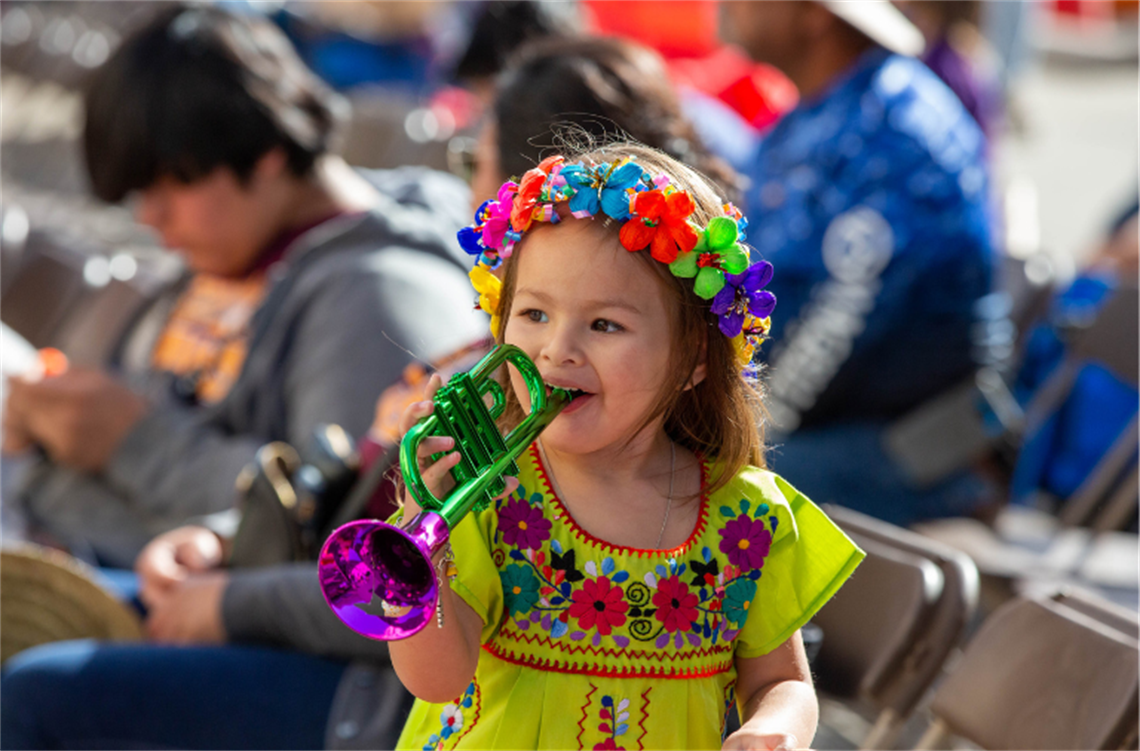 Child in colorful Fiesta attire playing a green and purple trumpet.