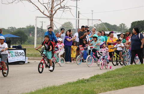 Children riding bikes past cheering families at an event, with a Vision Zero San Antonio table in the background.
