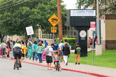 Students and parents walk and bike along a sidewalk toward an elementary school as a police officer rides behind them.