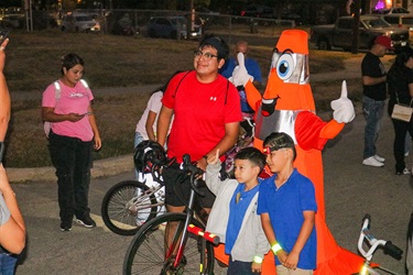 Children pose with a cyclist and a person in a traffic cone costume during an evening community biking event.