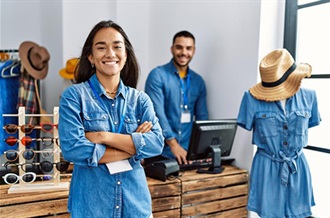A woman in a denim shirt poses in a store, with a friendly expression and a welcoming stance.