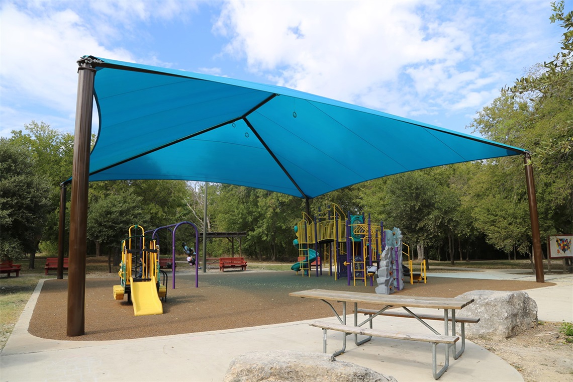 Colorful playground with yellow slides and climbing structures under a large blue shade canopy.