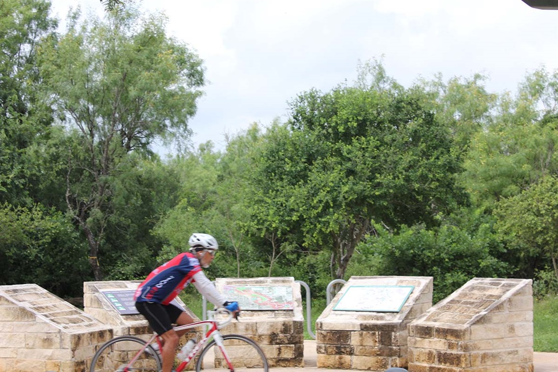 A man riding a bike past decorative stone-mounted park signs on a sunny day.