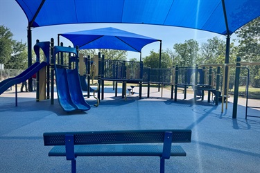 Playground with blue slides and shade canopies, seen from a nearby bench.