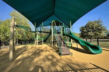 A playground with slides, climbing structures, and monkey bars, all under a large green shade canopy.